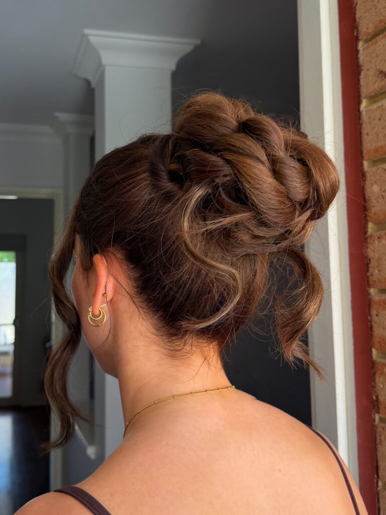 A woman with thick chestnut brown hair styled in a high twisted braided bun with wavy face-framing tendrils.