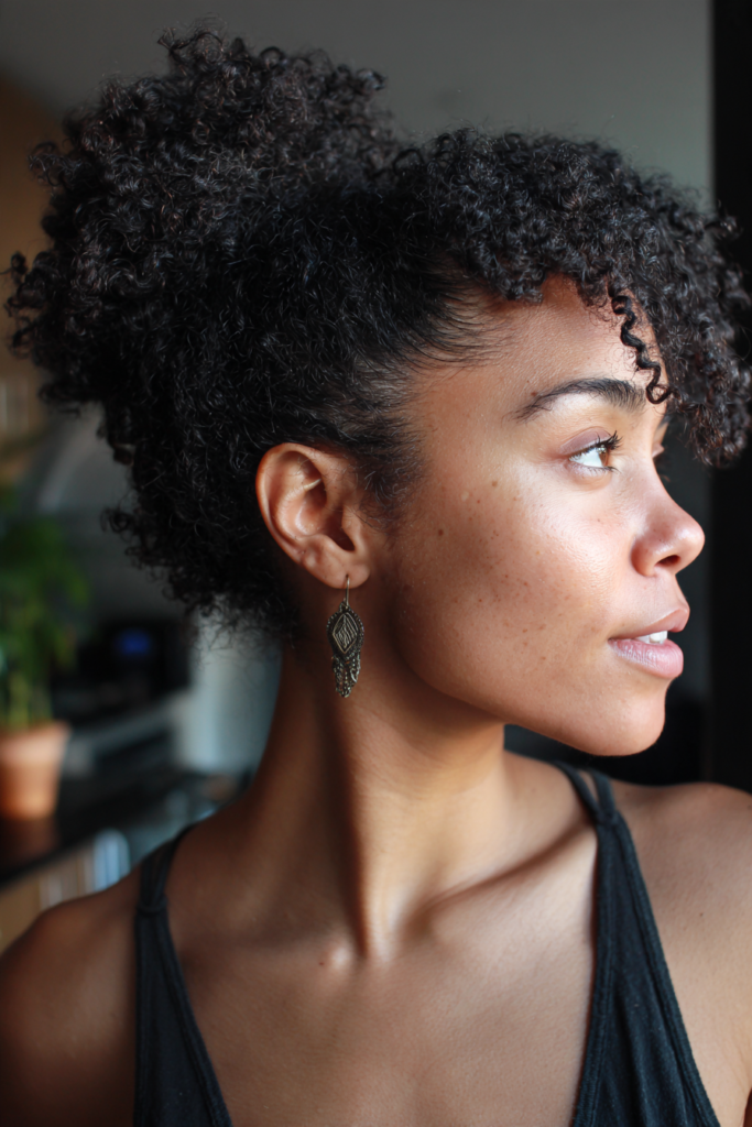A woman in profile wearing her dark coily hair in a high pineapple updo with textured curly bangs.
