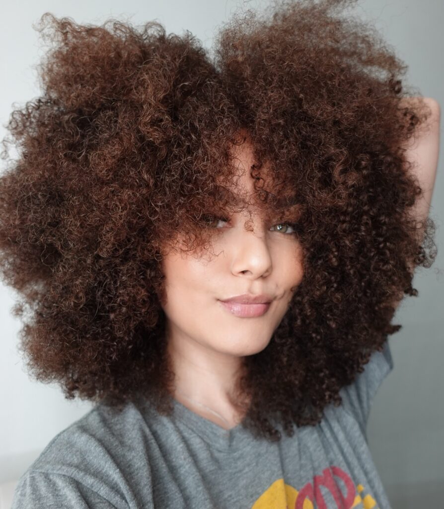 A woman with big voluminous chestnut brown coily hair and full curly bangs looking directly at the camera.