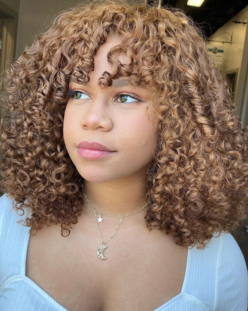 A woman with shoulder length honey blonde coily hair styled with curly bangs and layered volume.