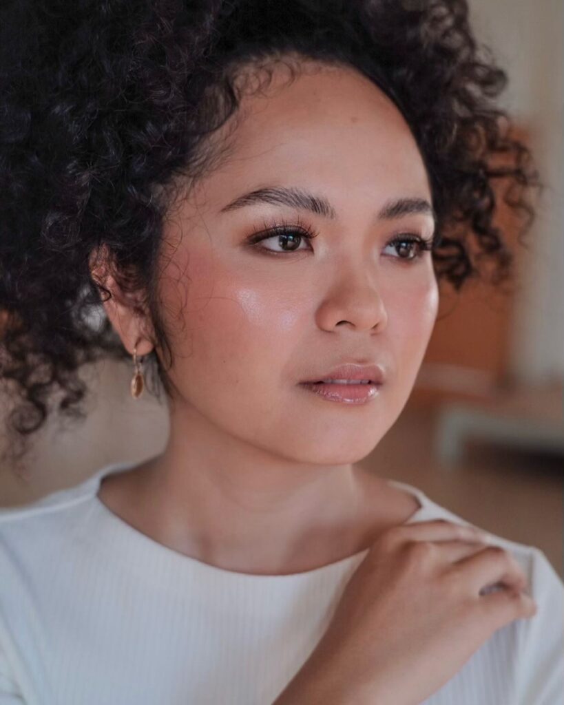 A woman with tight coily dark hair wearing a high curly puff updo with soft edges and a white top.