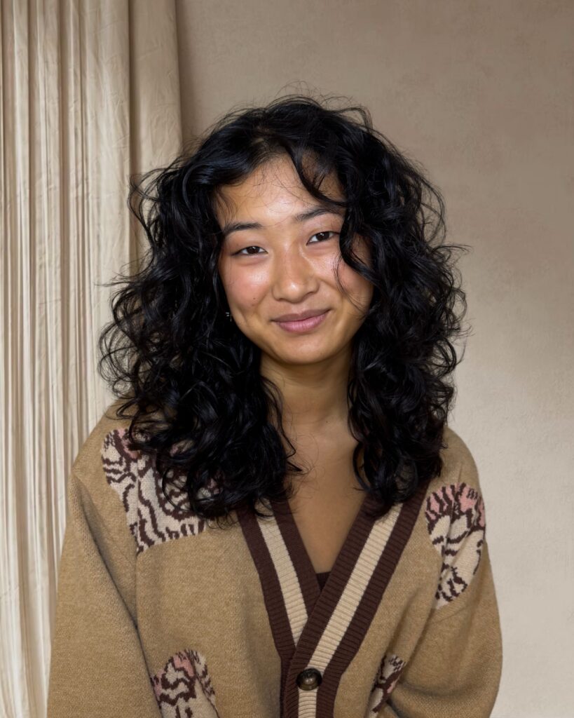 Woman with medium long jet black curly hair styled with layers, a soft side part and curly face framing pieces.