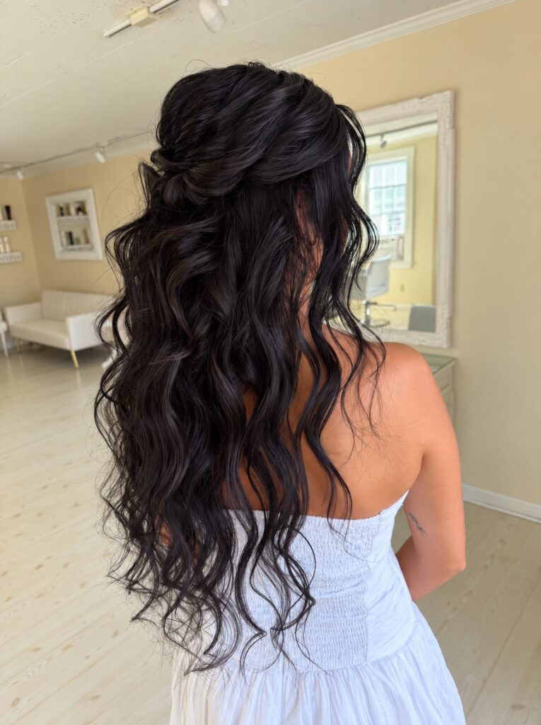 Back view of a woman with very long jet black wavy hair styled half up with a twisted knot at the crown.