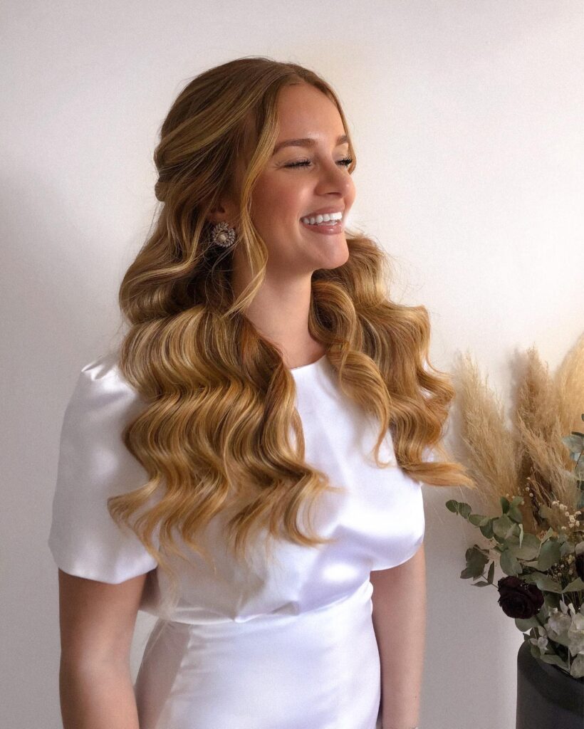 Side view of a smiling woman with very long golden honey blonde wavy hair styled half up with a center part and polished S waves.
