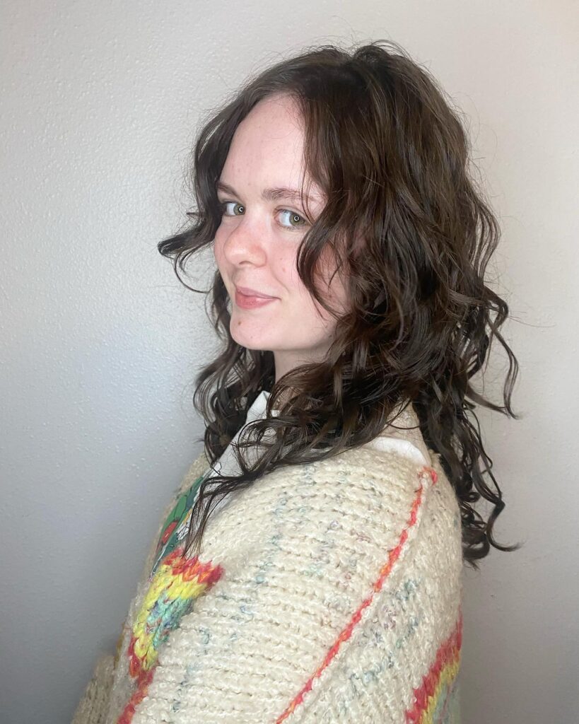 Side view of a woman with medium length dark brown wavy curly hair styled with layers, a center part and curled face framing pieces.