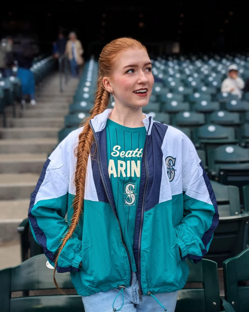 Woman with long copper red hair styled in a sleek slicked back fishtail braid, wearing a teal and navy Seattle Mariners jacket inside a stadium.