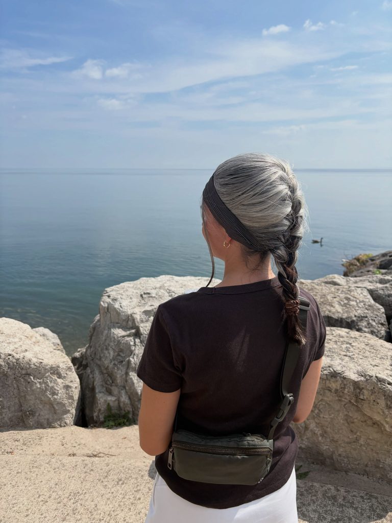Woman seen from behind with long silver gray hair styled in a French braid with a wide dark headband, standing by a lake with rocks.