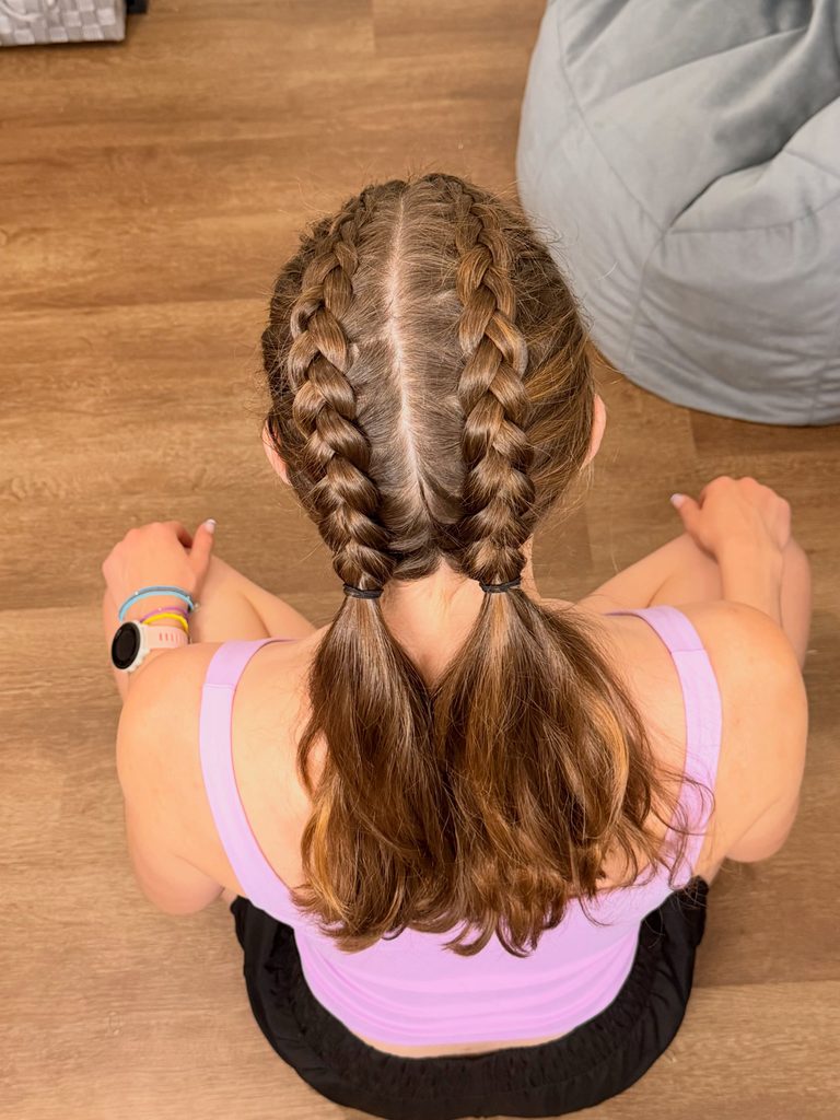 Woman seen from above and behind with medium length chestnut brown hair and caramel highlights styled in two Dutch braids that lead into low pigtails with a center part.