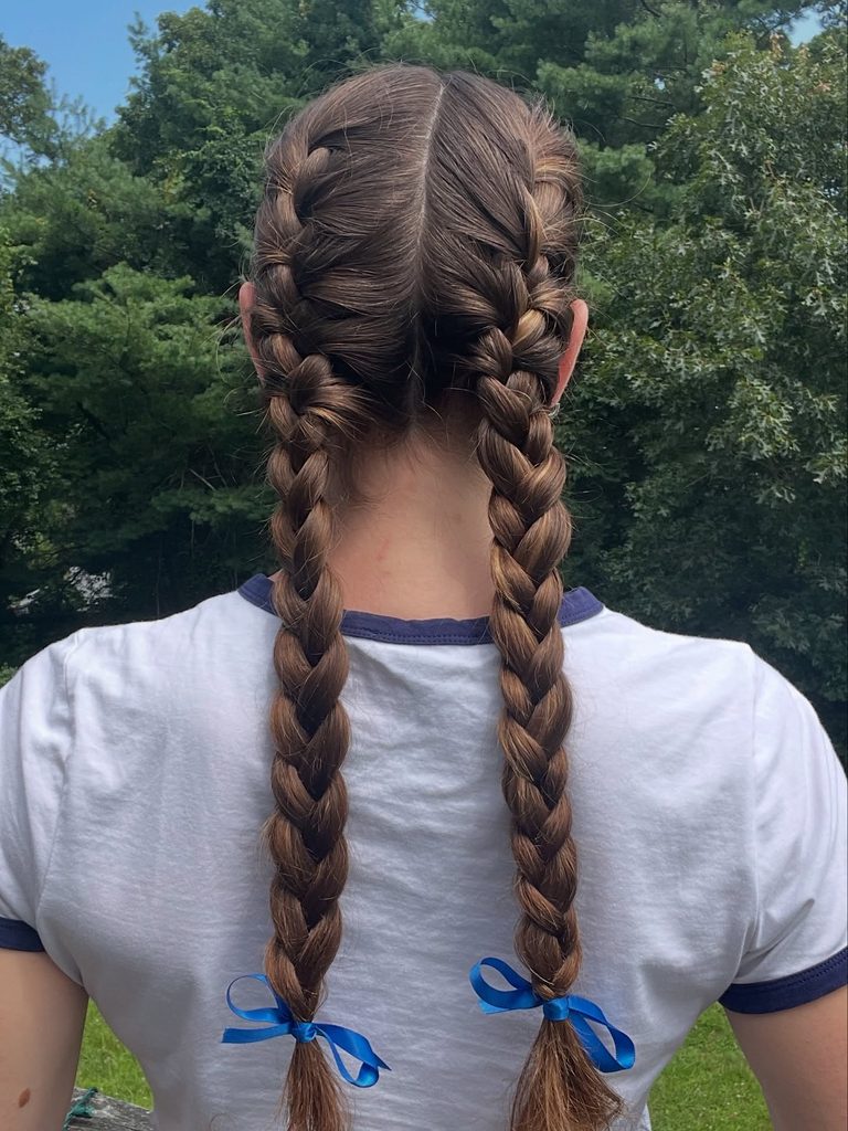 Woman seen from behind with long chestnut brown hair styled in two French braid pigtails with a center part and blue satin ribbon bows at the ends.