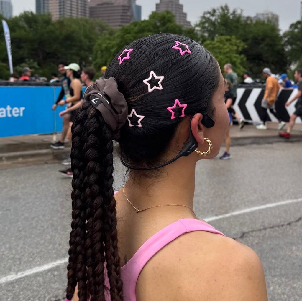 Woman seen from the side with long jet black hair in a sleek high ponytail of chunky braids with a brown scrunchie and pink star hair clips.