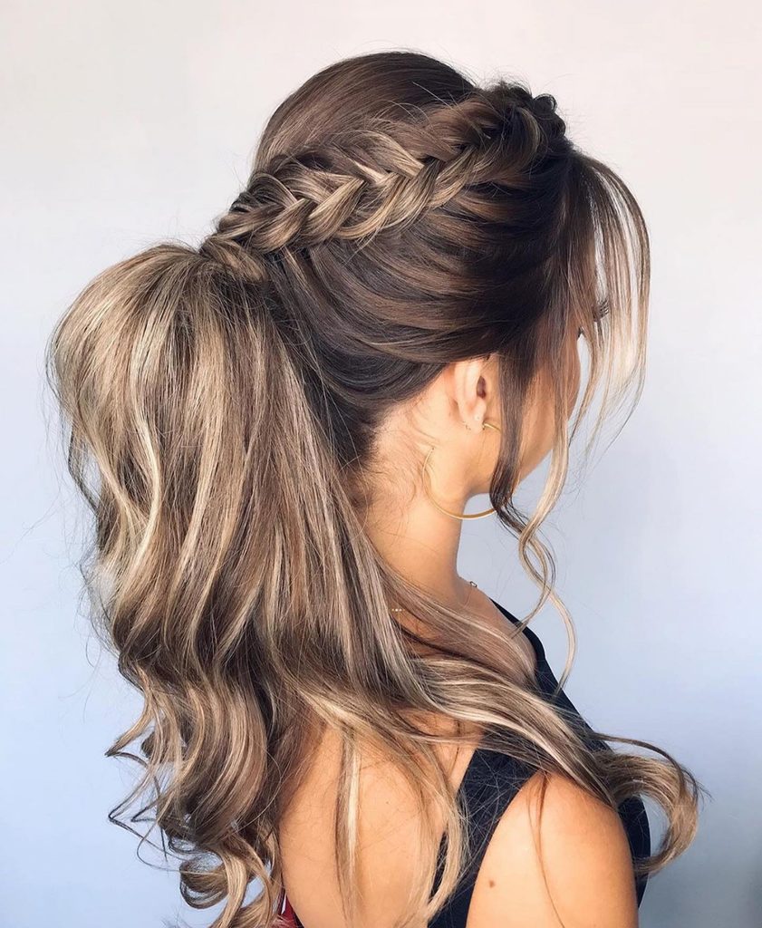 Woman seen from the side with long brunette hair and honey highlights styled in a braided crown that leads into a high wavy ponytail with loose face framing strands.
