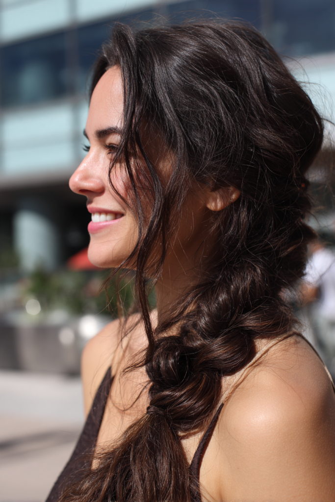 Side view of a smiling woman with long dark brown wavy hair styled in a loose, messy side bubble braid with face framing strands.