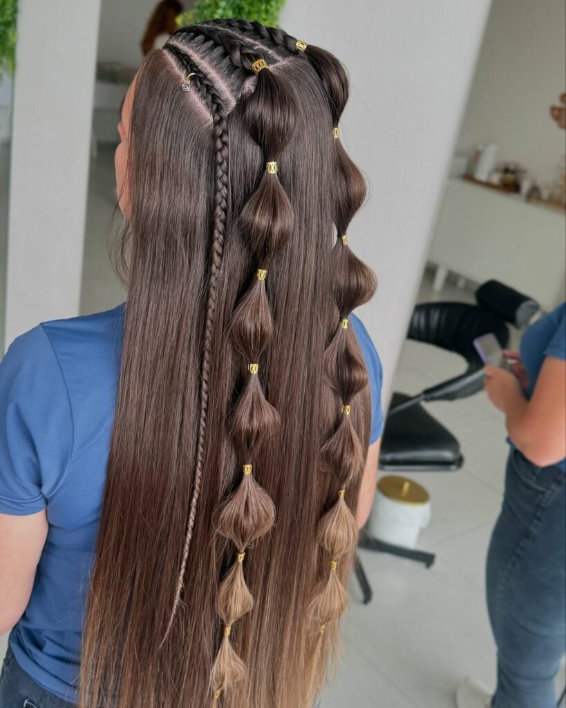 Back view of a woman with very long straight brunette hair with caramel ends styled with crown braids, a thin side braid, and two bubble braid sections with gold cuffs.