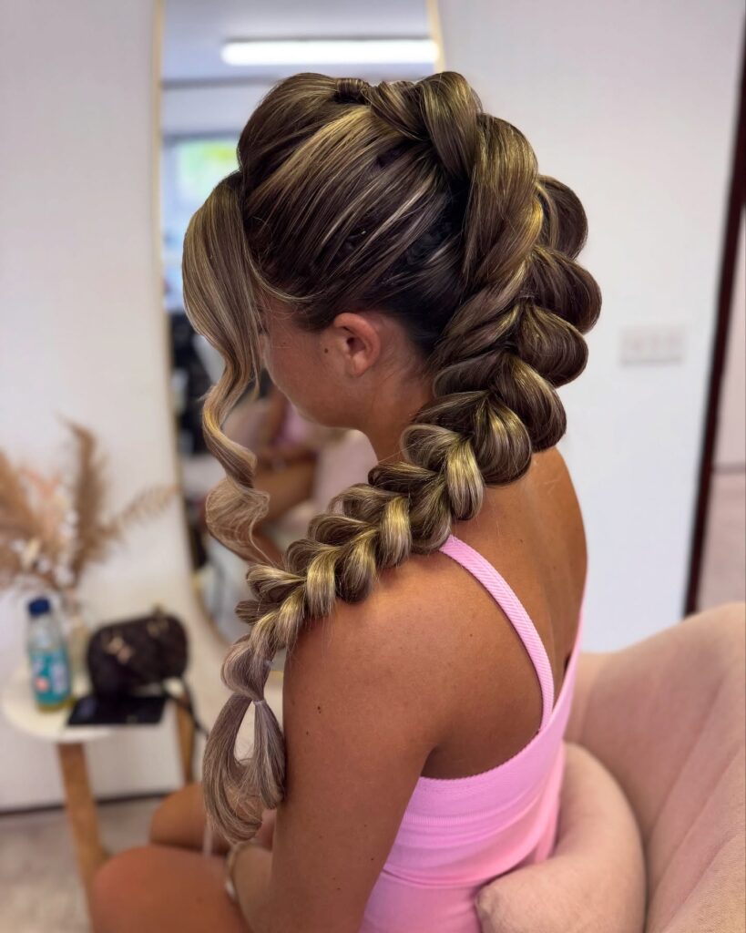 Side view of a woman with long brown hair and golden blonde highlights styled in a chunky side swept bubble braid with face framing curls and a clear elastic at the tip.