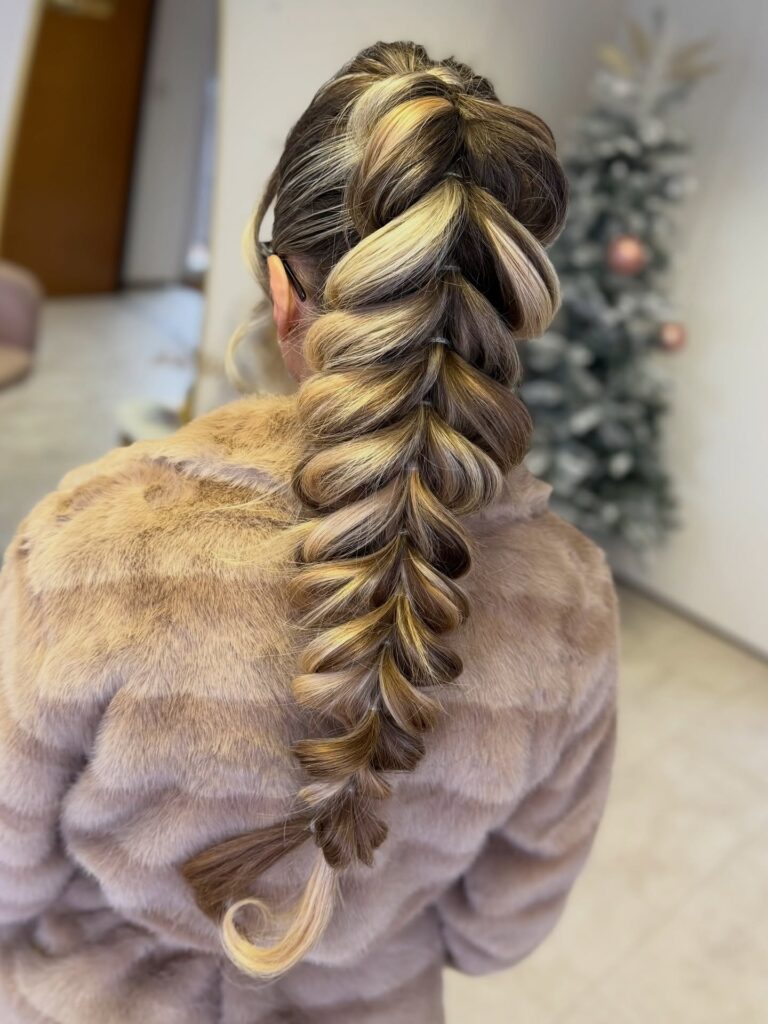 Back view of a woman with long brown hair and caramel blonde highlights styled in a high bubble braid with clear elastics and a curled blonde tip.