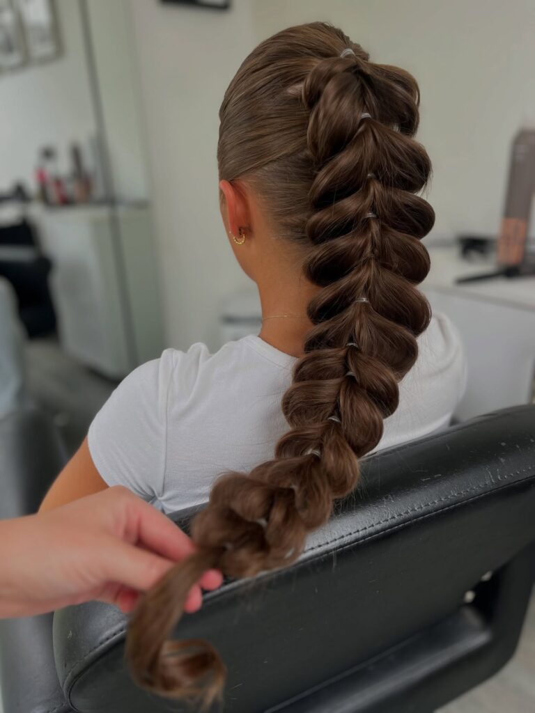Back view of a woman with long chestnut brown hair styled in a sleek high pull-through bubble braid with clear elastics and a tapered end.