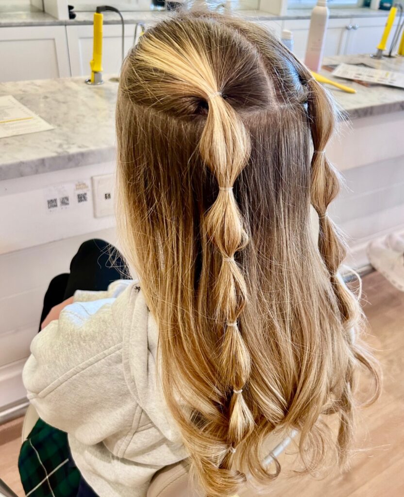 Back view of a woman with long brown to honey blonde ombre wavy hair styled half up with two small bubble braid sections secured with clear elastics.