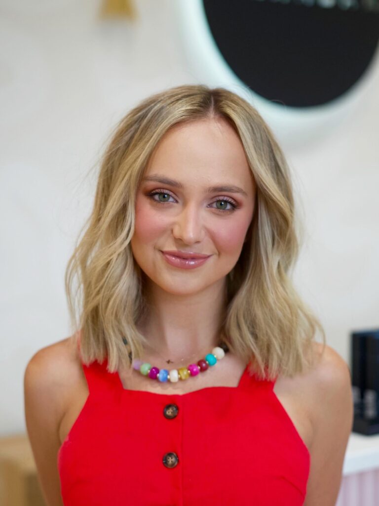 A smiling woman with a long blonde beachy wavy lob wears a red dress and beaded necklace.