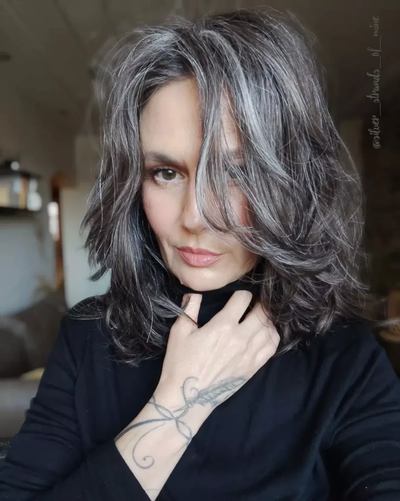 A woman with wavy shoulder-length salt and pepper hair and long face-framing layers poses with her hand near her chin.