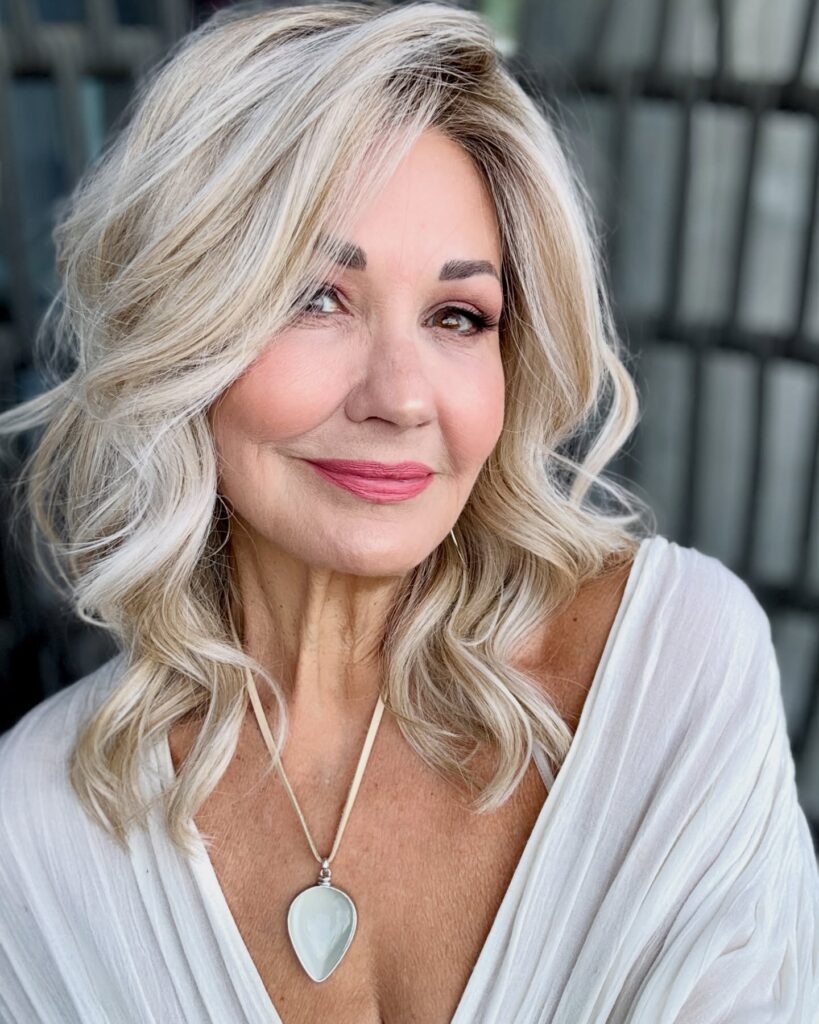 A smiling woman with voluminous platinum blonde curls and a deep side part wears a draped white top and pendant necklace.