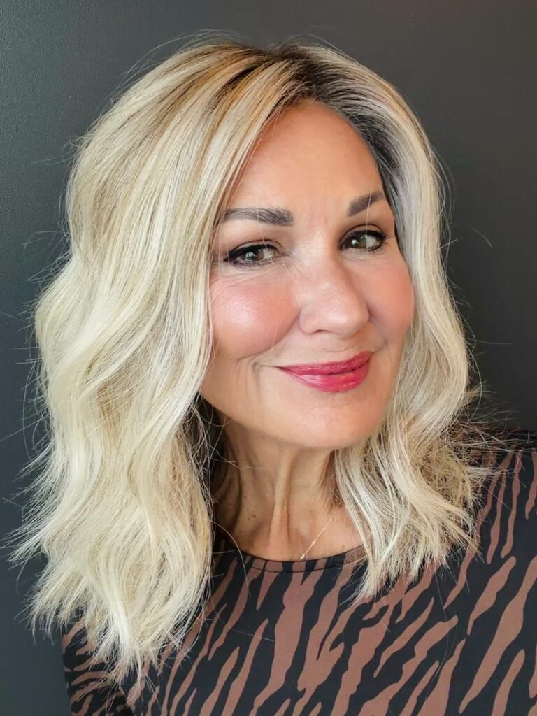 A woman with wavy shoulder-length icy blonde hair and a dark root shadow smiles while wearing a printed top.