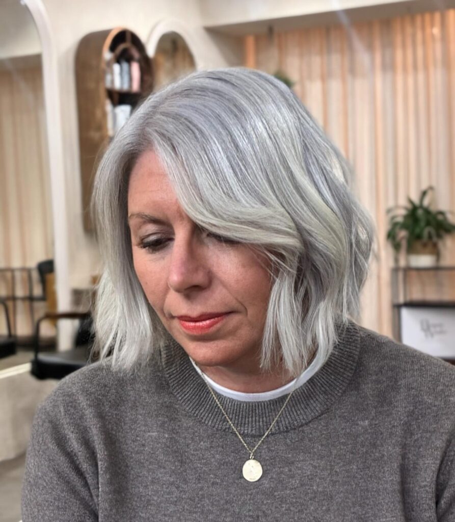 A woman with a shoulder-length silvery gray wavy lob looks down while wearing a gray sweater and pendant necklace.