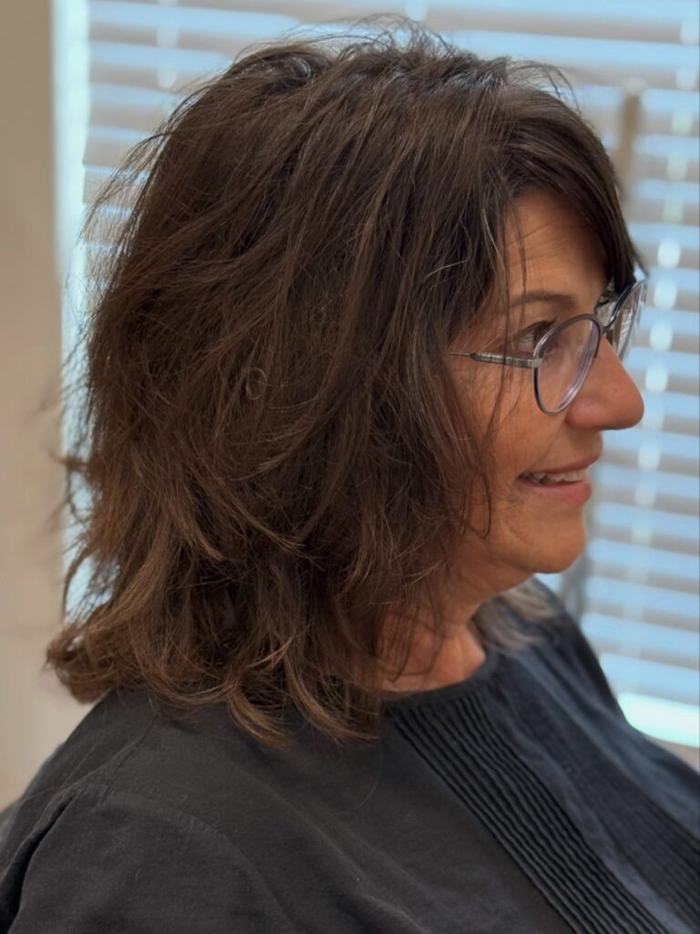 A smiling woman with a shaggy layered brunette hairstyle and wispy bangs wears glasses in profile view.