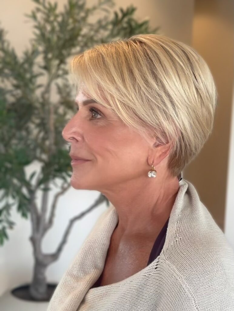 A woman with a short sleek golden blonde pixie cut and long side-swept bangs is shown in profile.