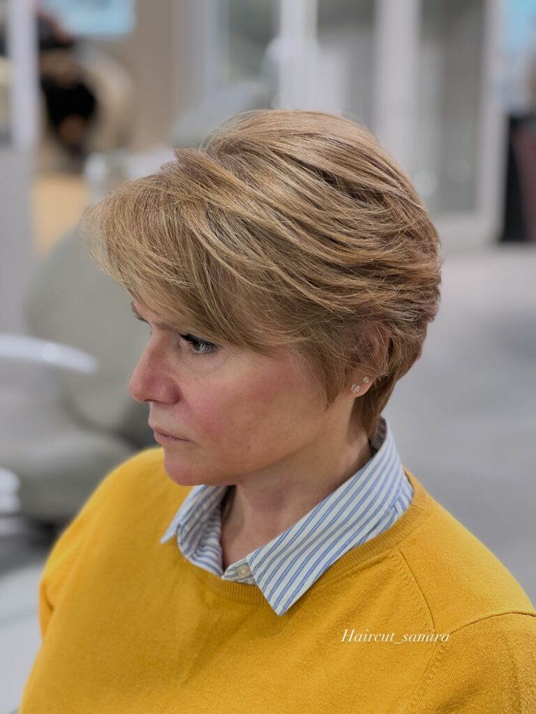 A woman with a feathered golden blonde pixie cut looks to the side wearing a yellow sweater.