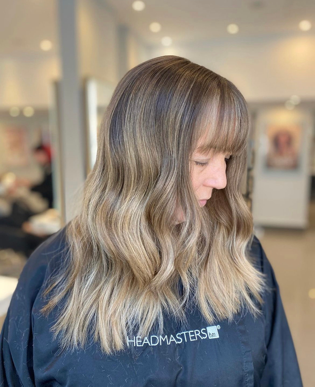 A woman with wavy balayage hair and a blunt fringe looks down while wearing a black salon cape.