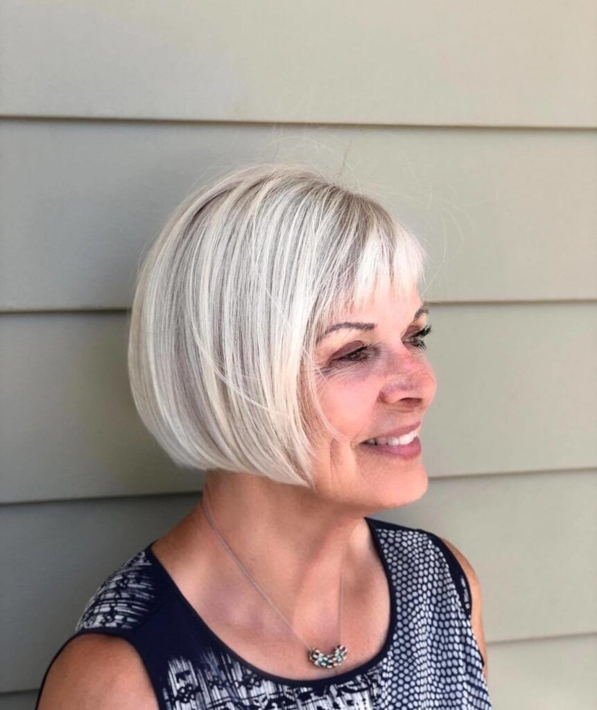 A smiling woman with a chin-length platinum blonde bob and soft wispy bangs stands against a light green paneled wall.