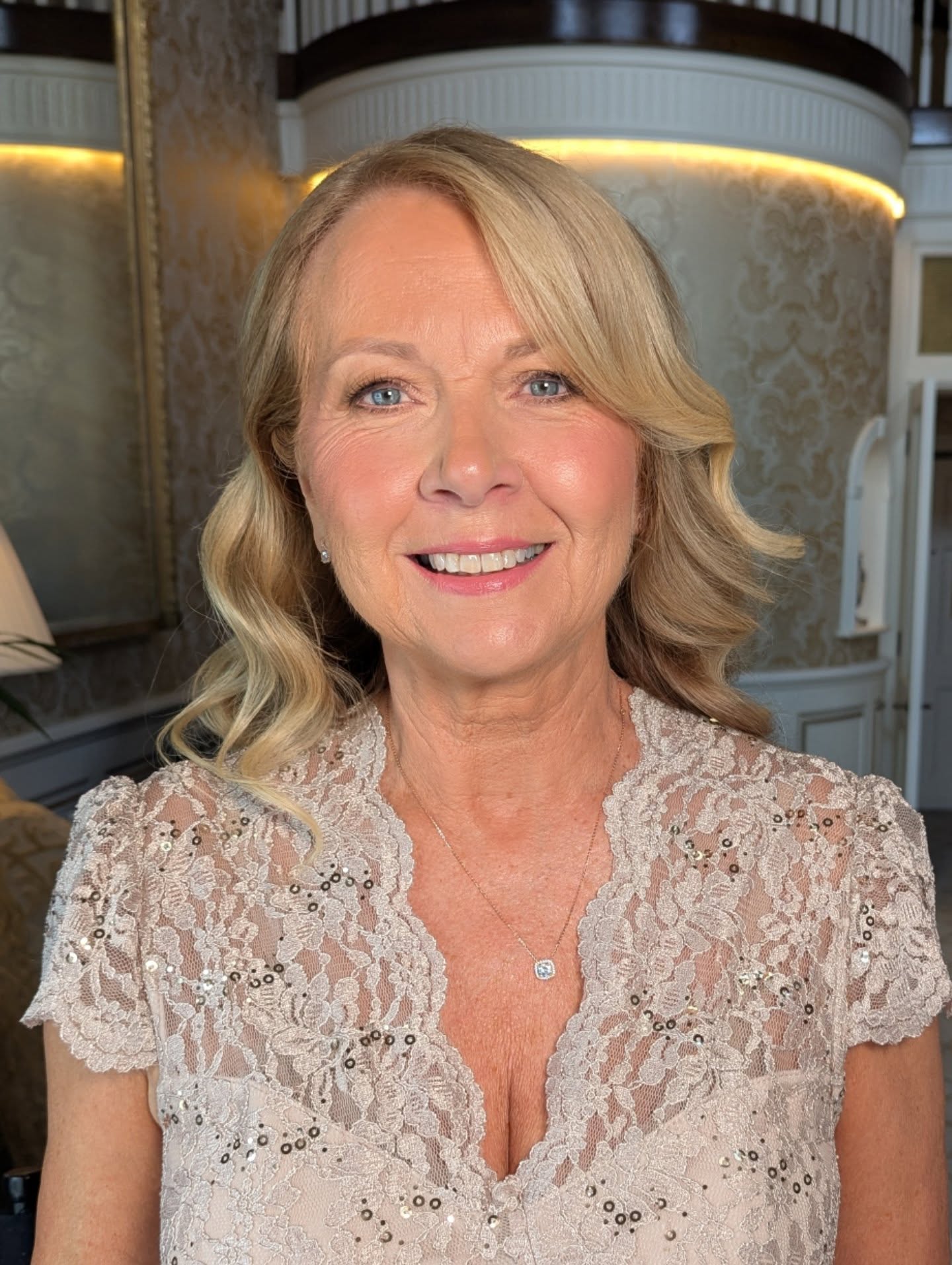 A smiling woman with medium-length honey blonde waves and a middle part wears a lace sequined top and pendant necklace.