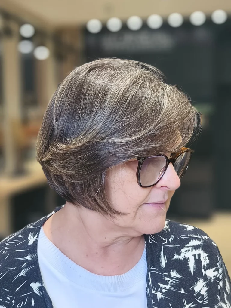 A woman with a feathered salt and pepper bob and curved ends wears glasses and a patterned cardigan.