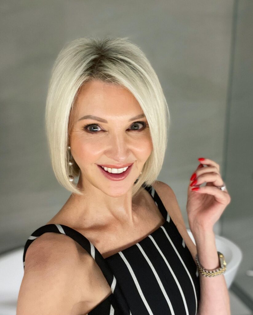 A smiling woman with a sleek platinum blonde chin-length bob wears a striped top and red nail polish.