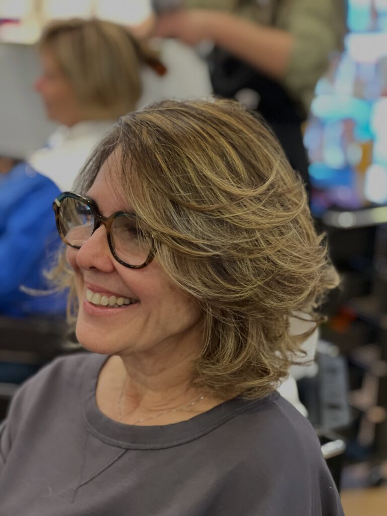 A smiling woman with a voluminous shoulder-length bob featuring flipped-out ends and golden highlights wears glasses.