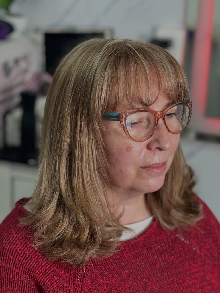 A woman with shoulder-length wavy blonde hair and full blunt bangs wears glasses and a red sweater.