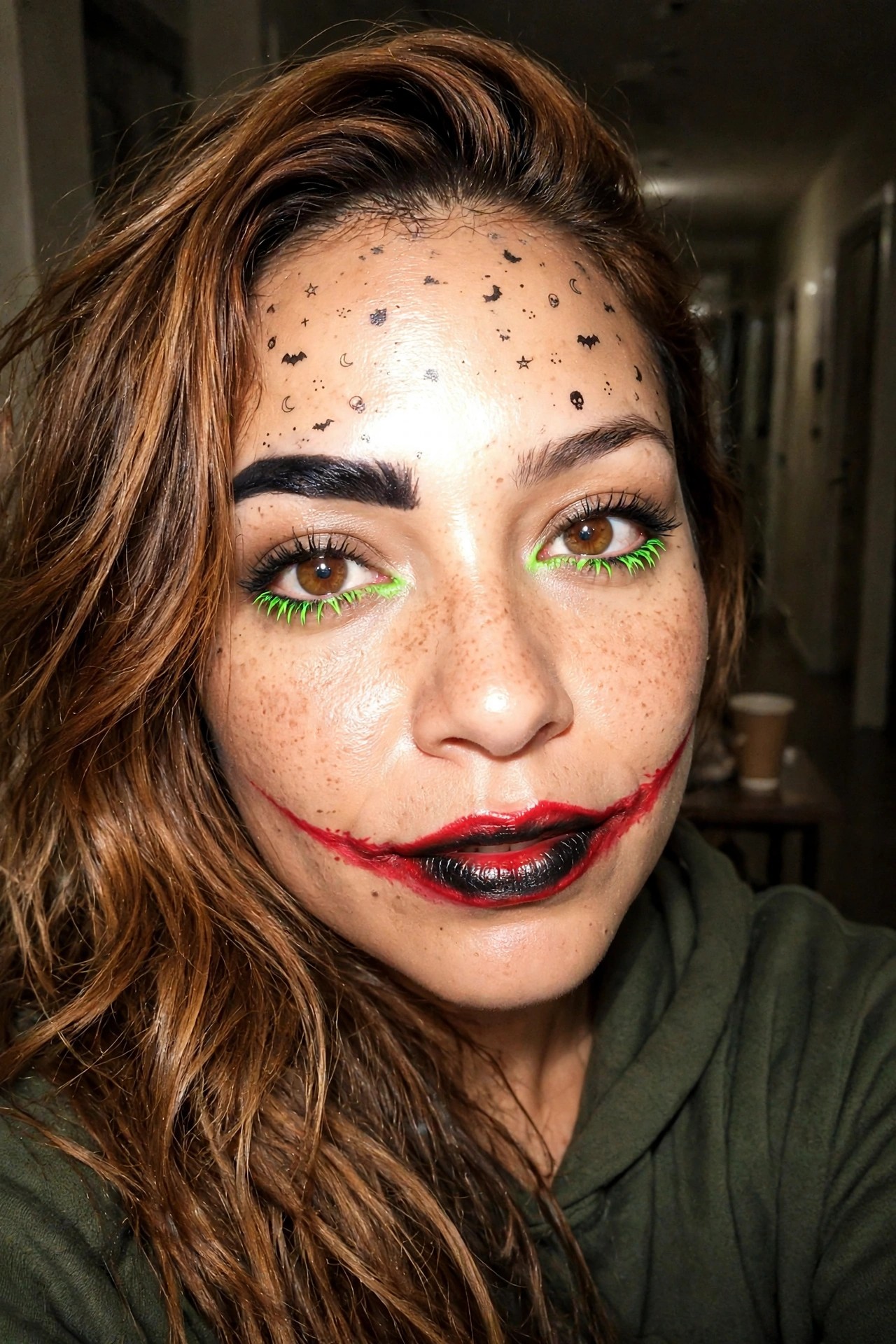 Close up of Joker style makeup with red smile extending from the mouth, black lips, neon green eyelashes, and small black Halloween symbols on the forehead.