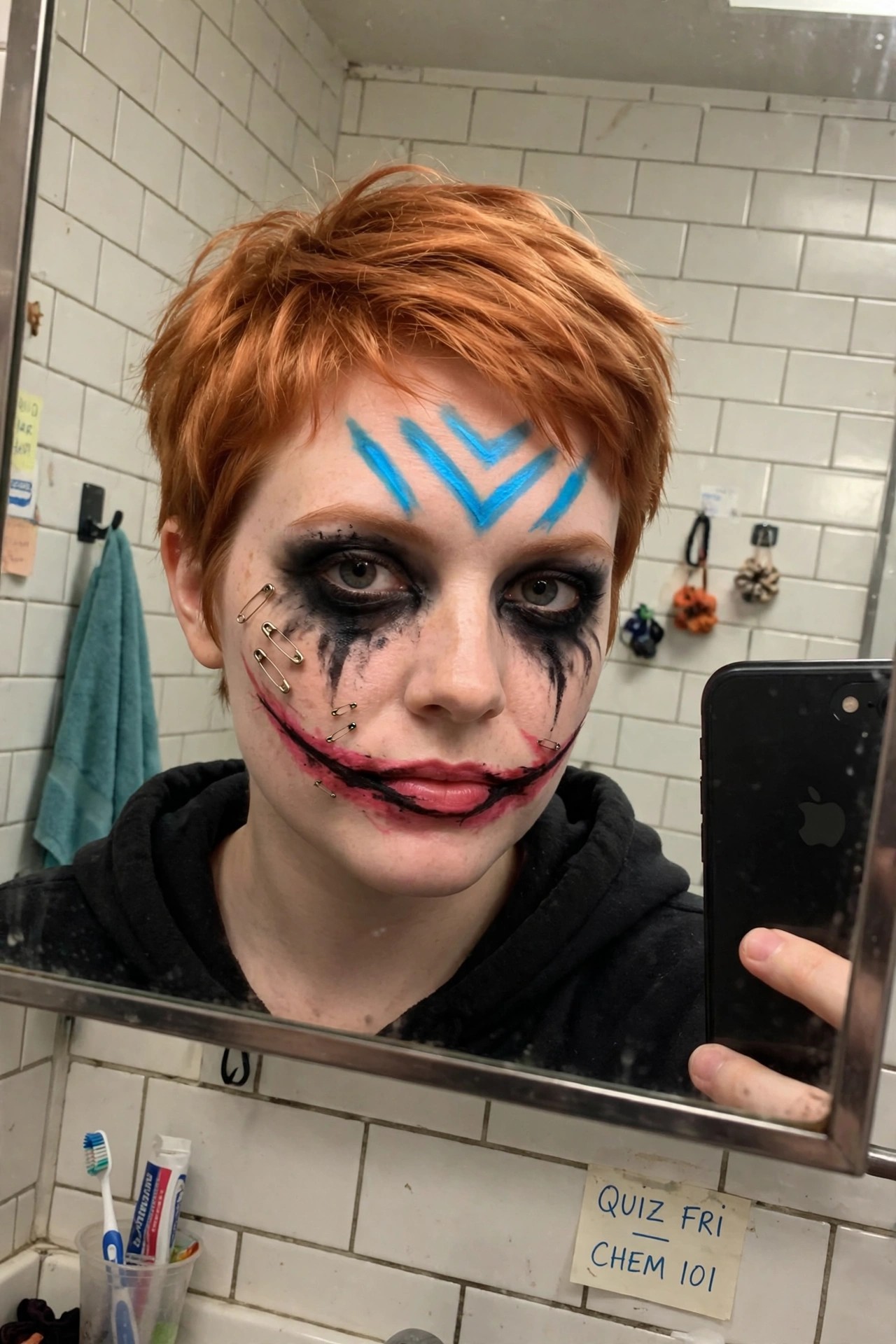 Close up of Joker style makeup featuring blue chevron shapes on the forehead, heavy smudged black eye makeup with downward streaks, red and black painted grin, and small safety pins placed along the cheek and jaw.