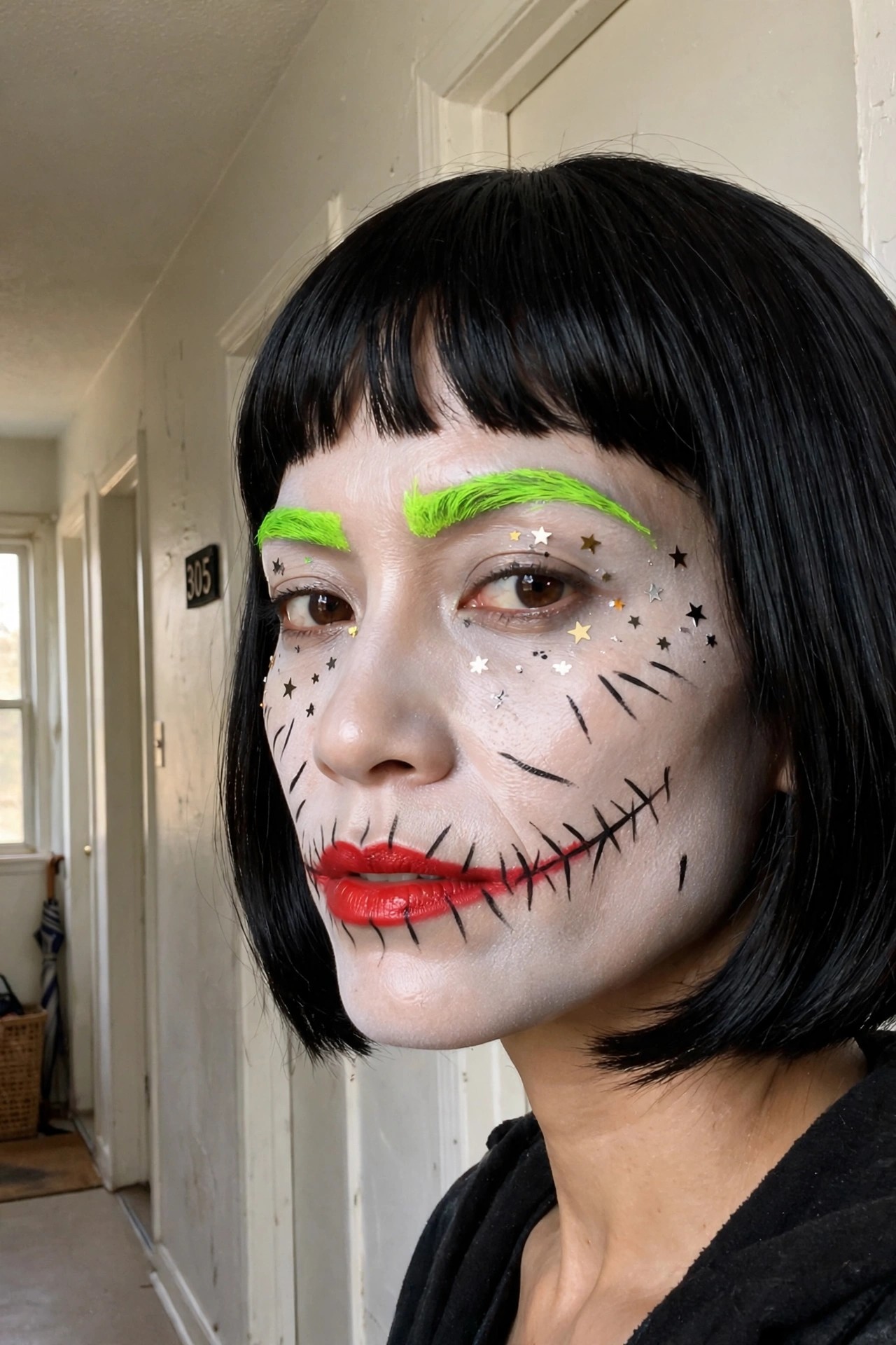 Close up of Joker style Halloween makeup with white face paint, neon green eyebrows, red lips, black stitch lines around the mouth and cheeks, and glitter star accents near the eyes.