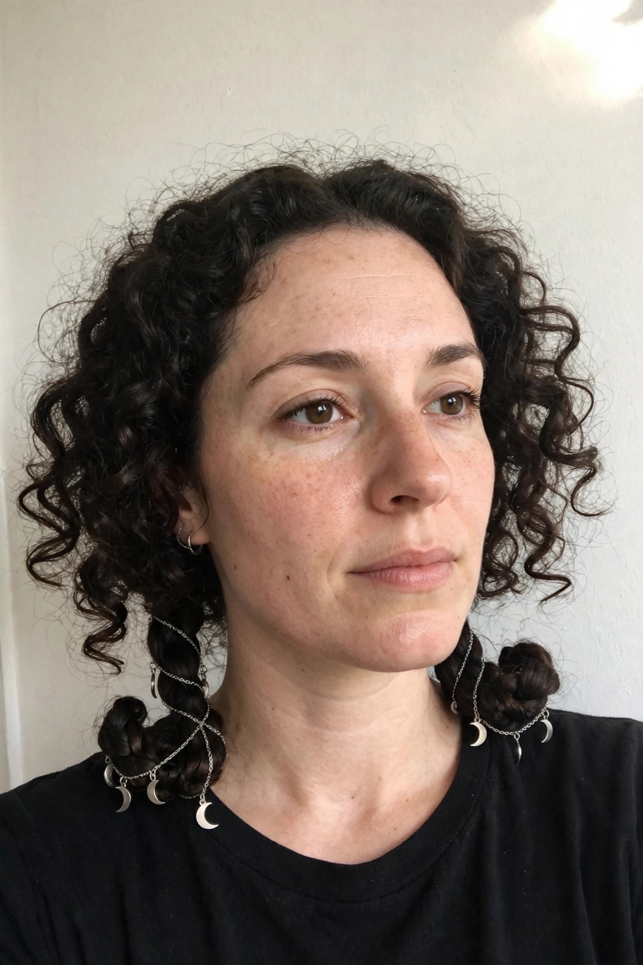 Dark curly hair styled in two low braided sections with silver chains and crescent moon charms dangling from the braids.