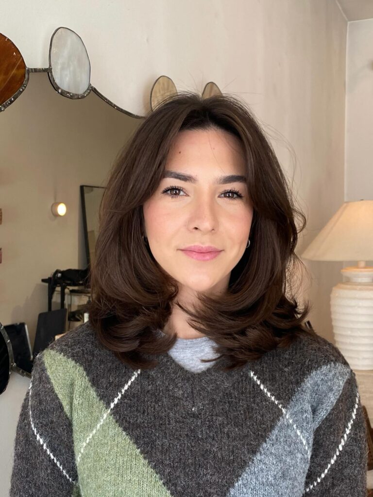 A woman with shoulder-length, voluminous brown hair styled in soft flipped layers with a center part and face-framing pieces.
