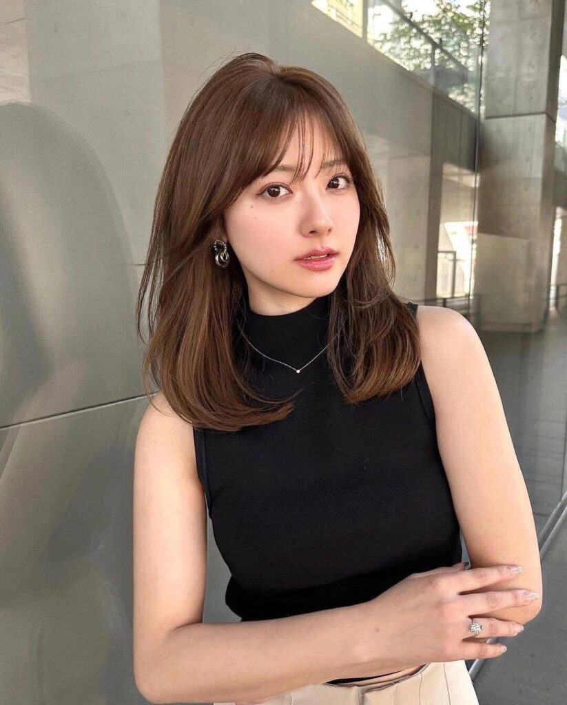 A woman with shoulder-length brown hair featuring soft wispy curtain bangs and gentle waves at the ends.