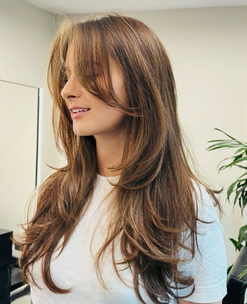 A woman with long, layered bronde hair featuring curtain bangs and warm caramel highlights throughout.