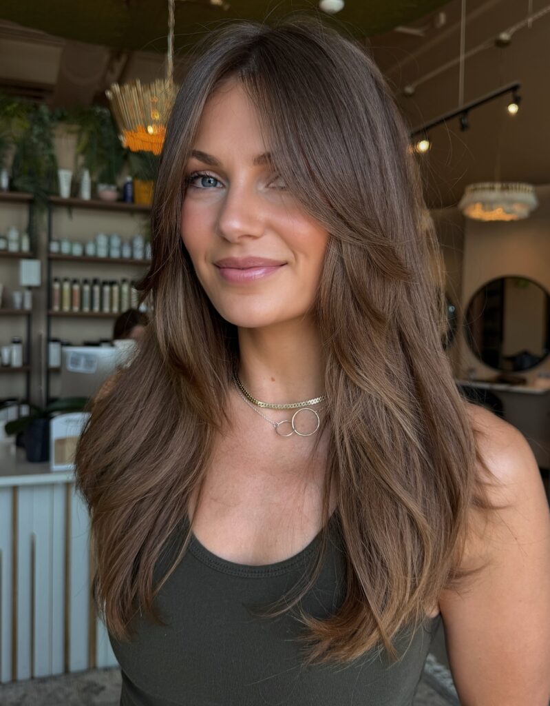 A woman with long, layered brunette hair featuring soft waves, subtle highlights, and voluminous bounce throughout.