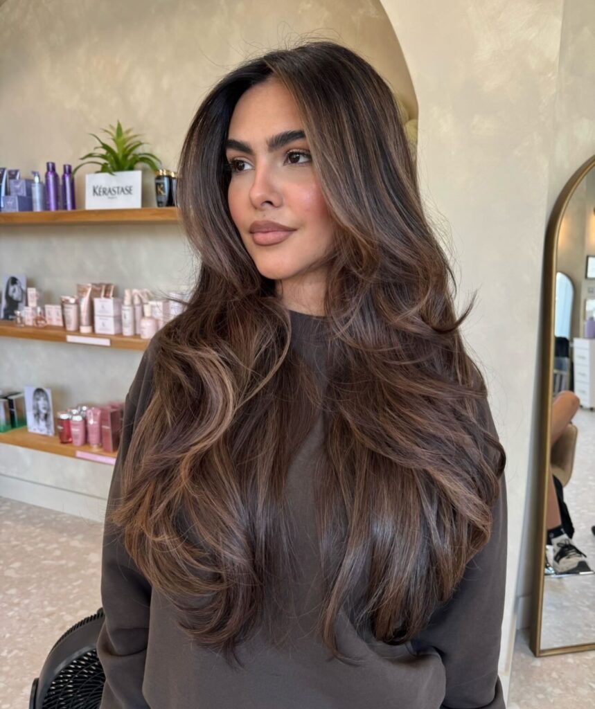  A woman with long, wavy chestnut brown hair featuring caramel balayage and voluminous layers throughout.