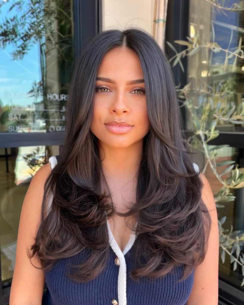 A woman with long, sleek dark brown hair featuring a center part and soft flipped-out layers at the ends.