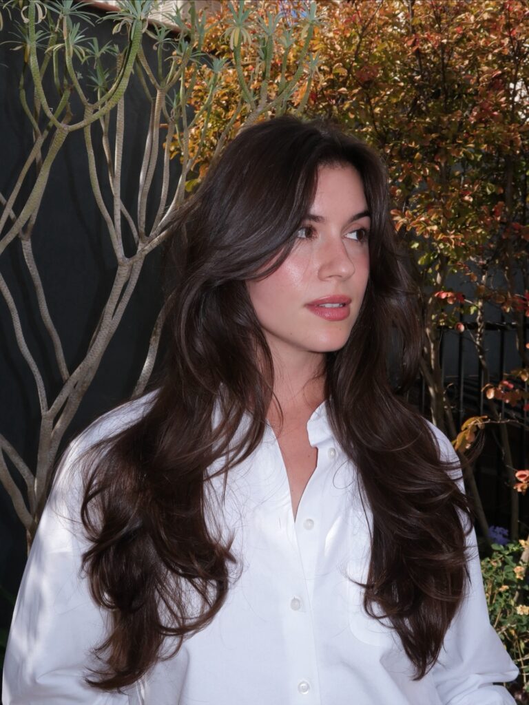 A woman with long, wavy deep brunette hair featuring voluminous face-framing layers and soft curled ends.