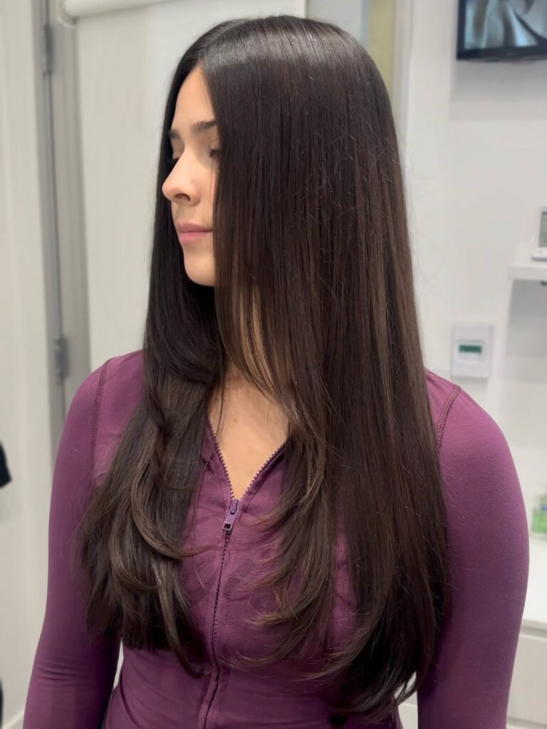 A woman with long, sleek dark brunette hair featuring a deep side part and subtle flipped ends with caramel highlights.