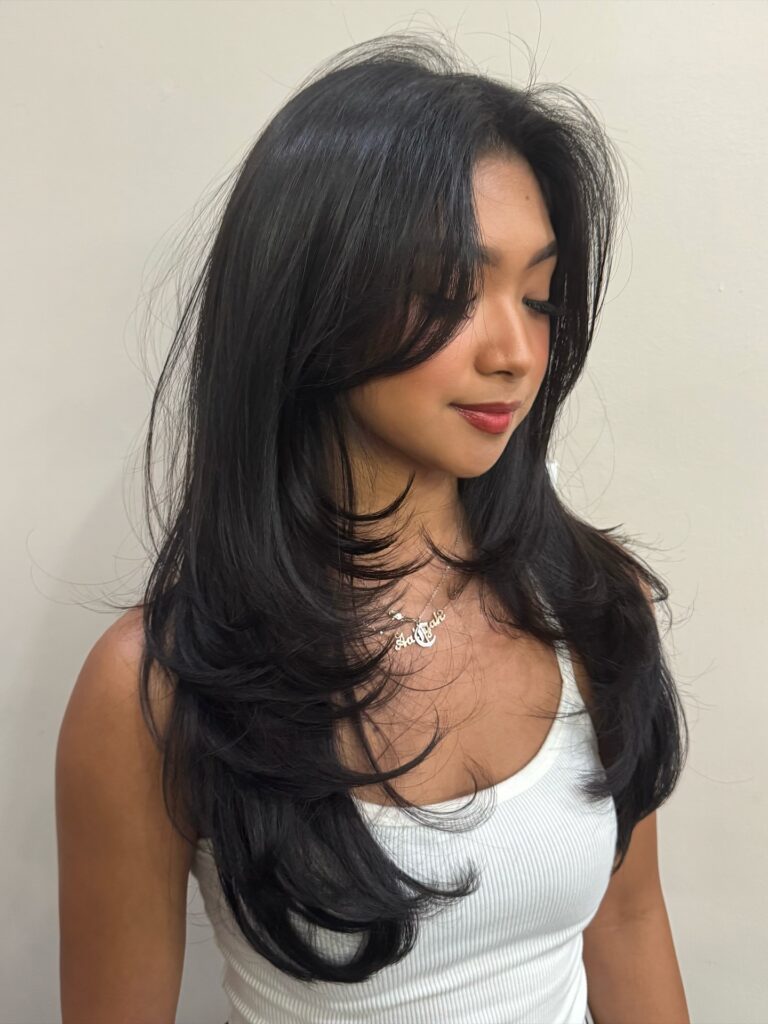 A woman with long, jet black layered hair featuring soft curled-under ends framing the face and neckline.