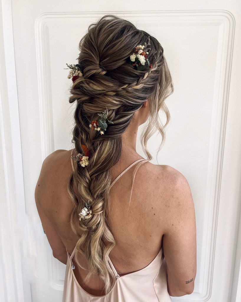 Back view of a woman with long brunette hair and blonde highlights styled in a twisted boho braid decorated with small dried flower hair pins.
